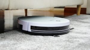 Robotstofzuiger reinigt zelfstandig onder een bank op een grijs tapijtt, automatisering in de schoonmaak